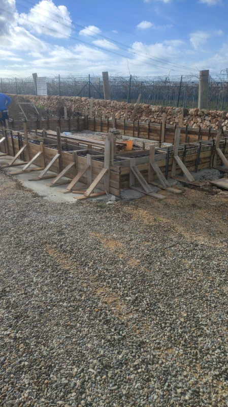 Cantiere edile all'aperto con lunghe casseforme per barre d'armatura e supporti in legno su ghiaia sotto un cielo nuvoloso.