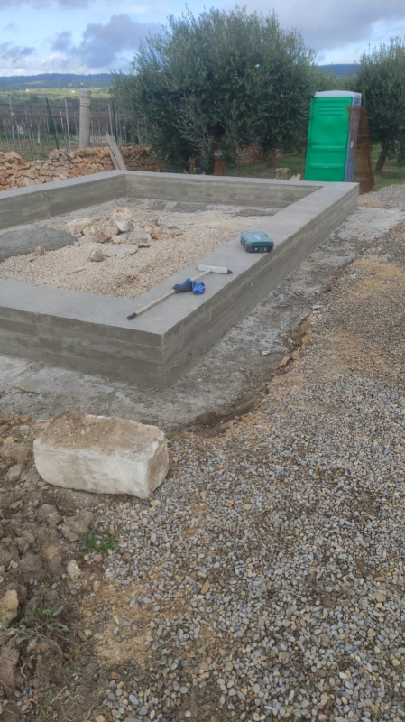 Soletta di fondazione in cemento in costruzione su un terreno ghiaioso, con una vasca verde sullo sfondo.