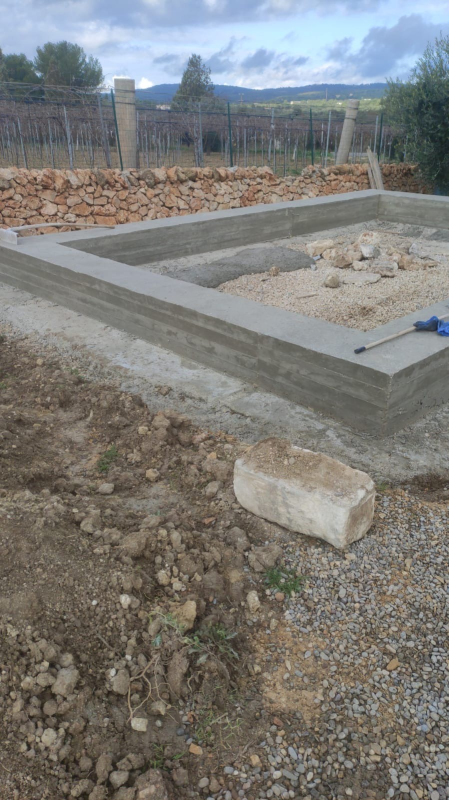 Impronta di fondazione in cemento su un lotto di terreno sterrato, con un bordo in pietra e colline boscose sullo sfondo.