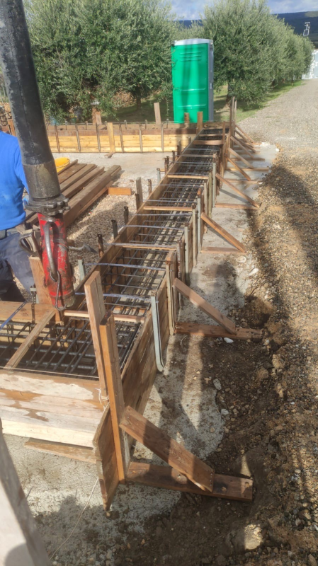 Fondamenta in ferro in costruzione accanto a un sentiero sterrato, con casseforme in legno e un bagno chimico verde sullo sfondo.