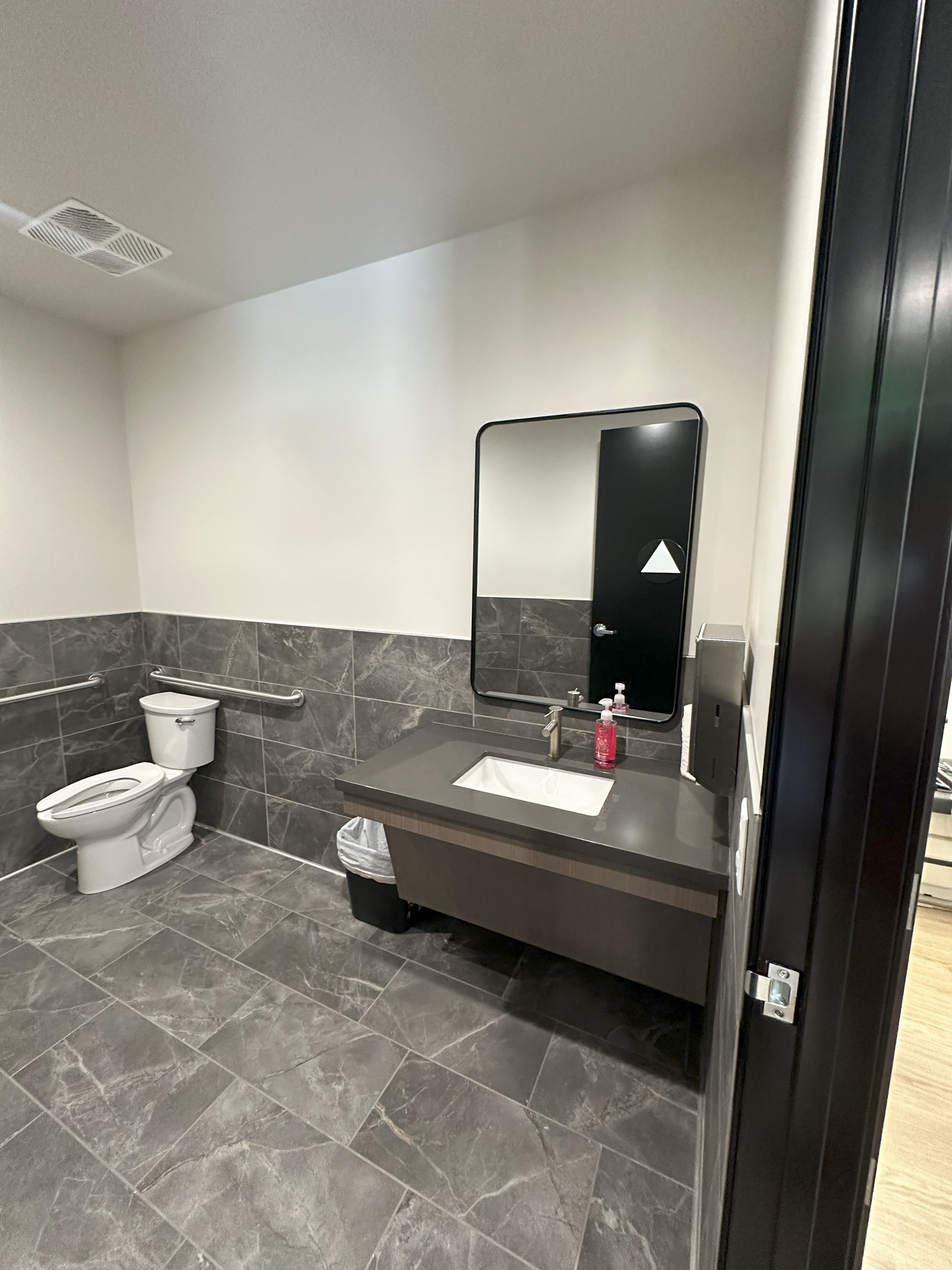 Bathroom with gray tile, toilet, sink, and large mirror. Handrails present.
