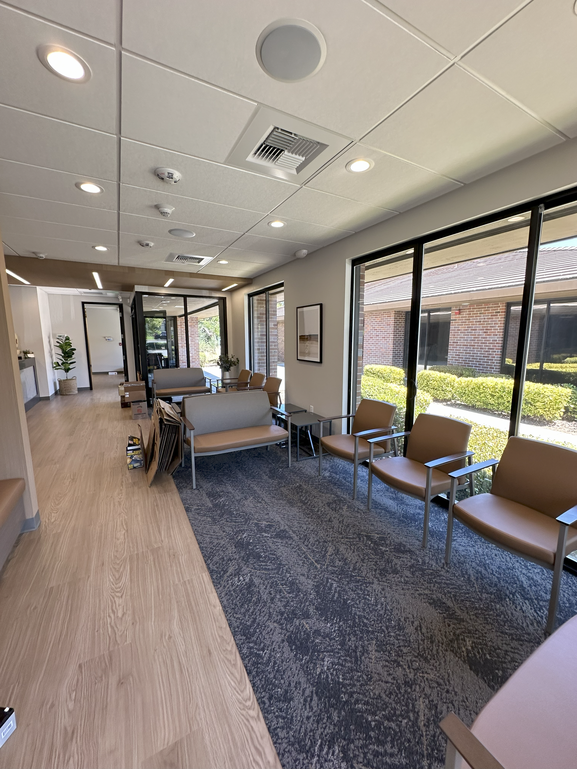 A modern medical waiting room with rows of chairs, a gray rug, and large windows overlooking a courtyard.