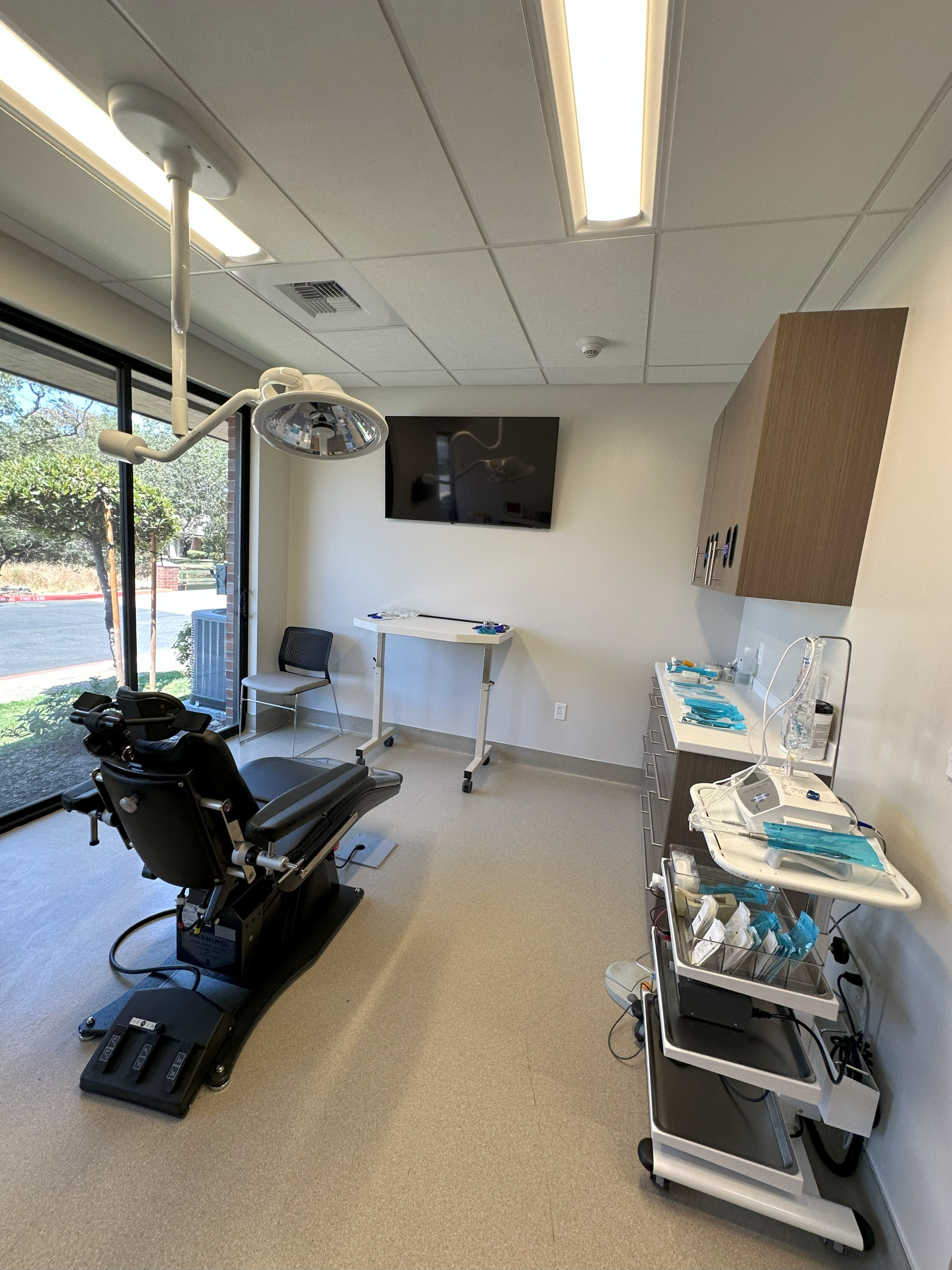 A dental examination room featuring a dental chair, equipment carts, overhead light, and a wall-mounted monitor.