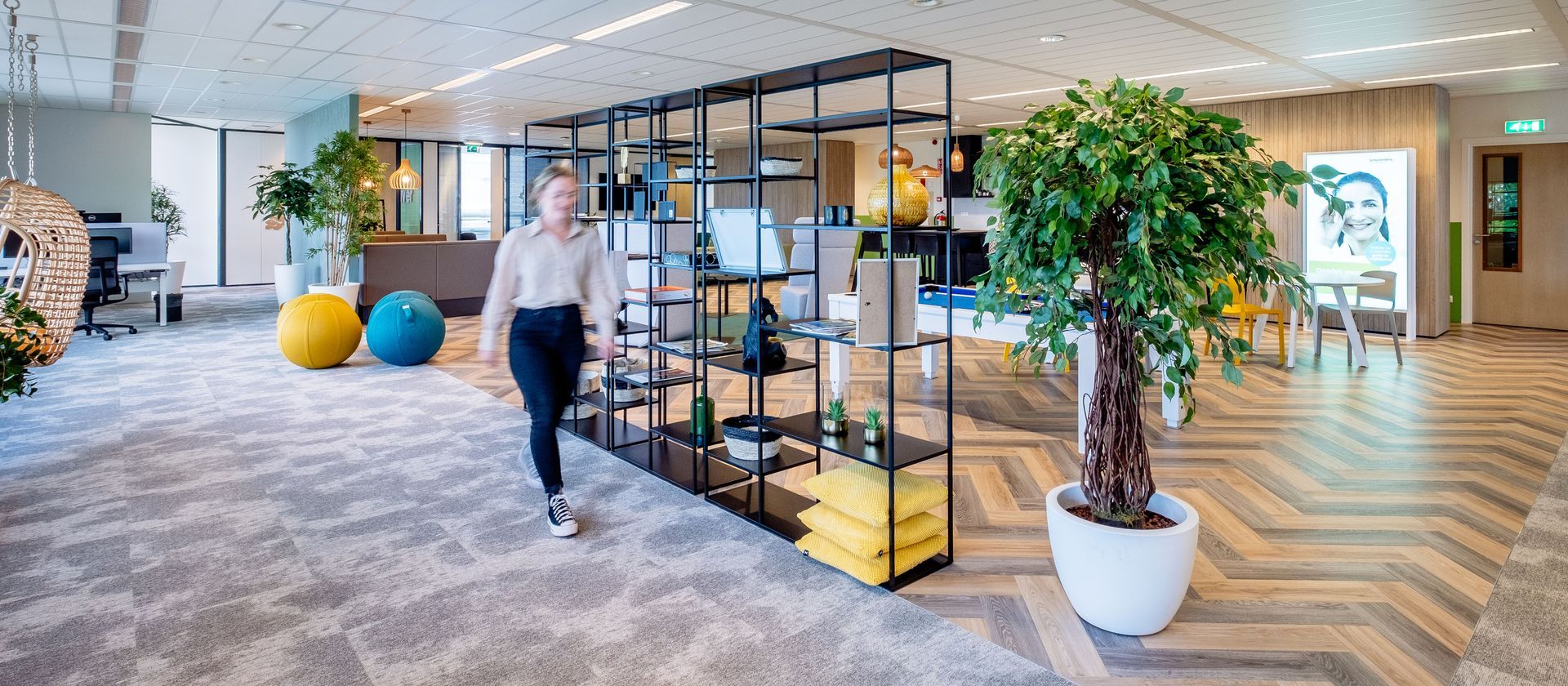 Bright office lounge with carpeted seating area, shelves, potted tree, and a person walking through the space.