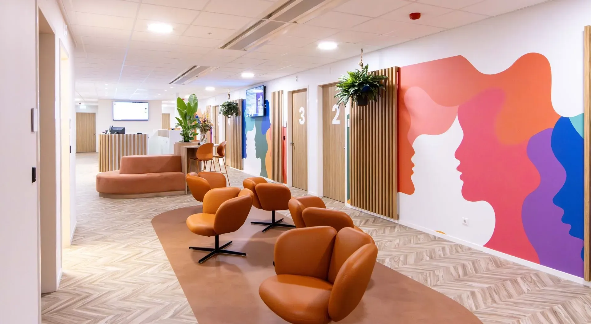 Moderne kantoorhal met oranje loungestoelen, potplanten en kleurrijke abstracte muurdecoratie.