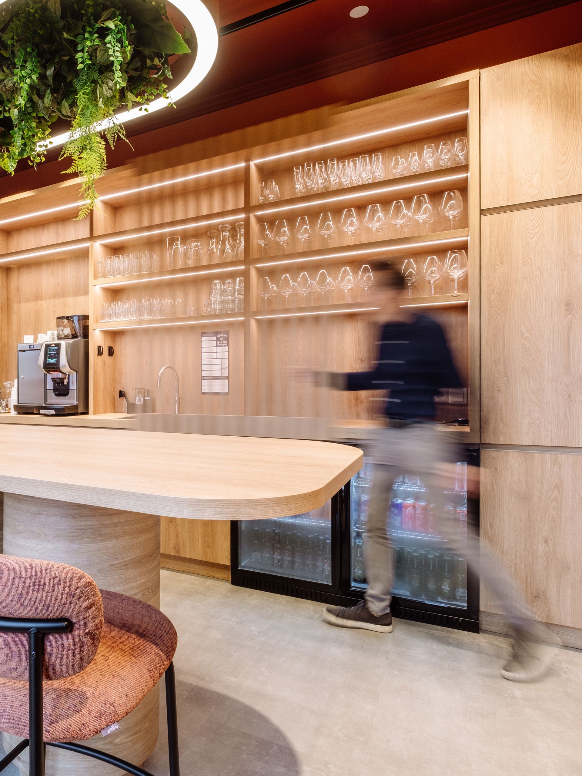 Modern café-interieur met glazen planken, een grote tafel en een wazige figuur die langs de toonbank loopt in kantoor