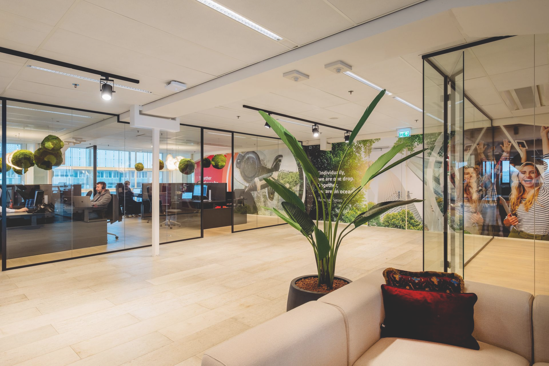 Moderne kantoorlounge met glazen wanden, beige bank en een grote potplant naast een zittende persoon op de achtergrond.