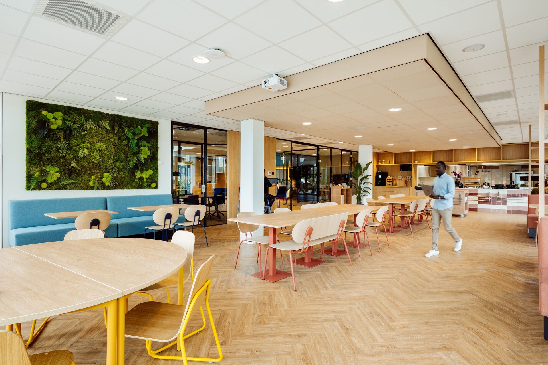 Modern café-interieur met houten tafels, blauwe stoelen, groene muurdecoratie en een voorbijlopende persoon.interieurontwerp door KuóStudio