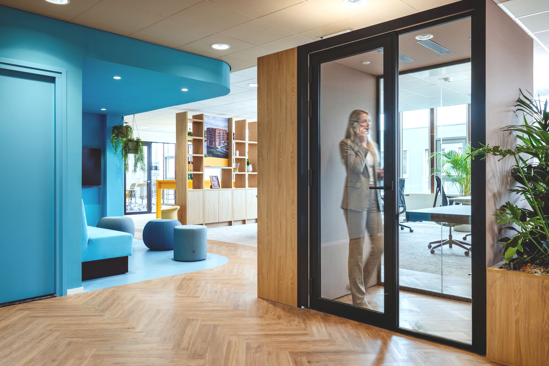 Moderne kantoorlobby met glazen vergaderruimte, blauwe zitmeubels, houten accenten en een persoon in de telefooncel.