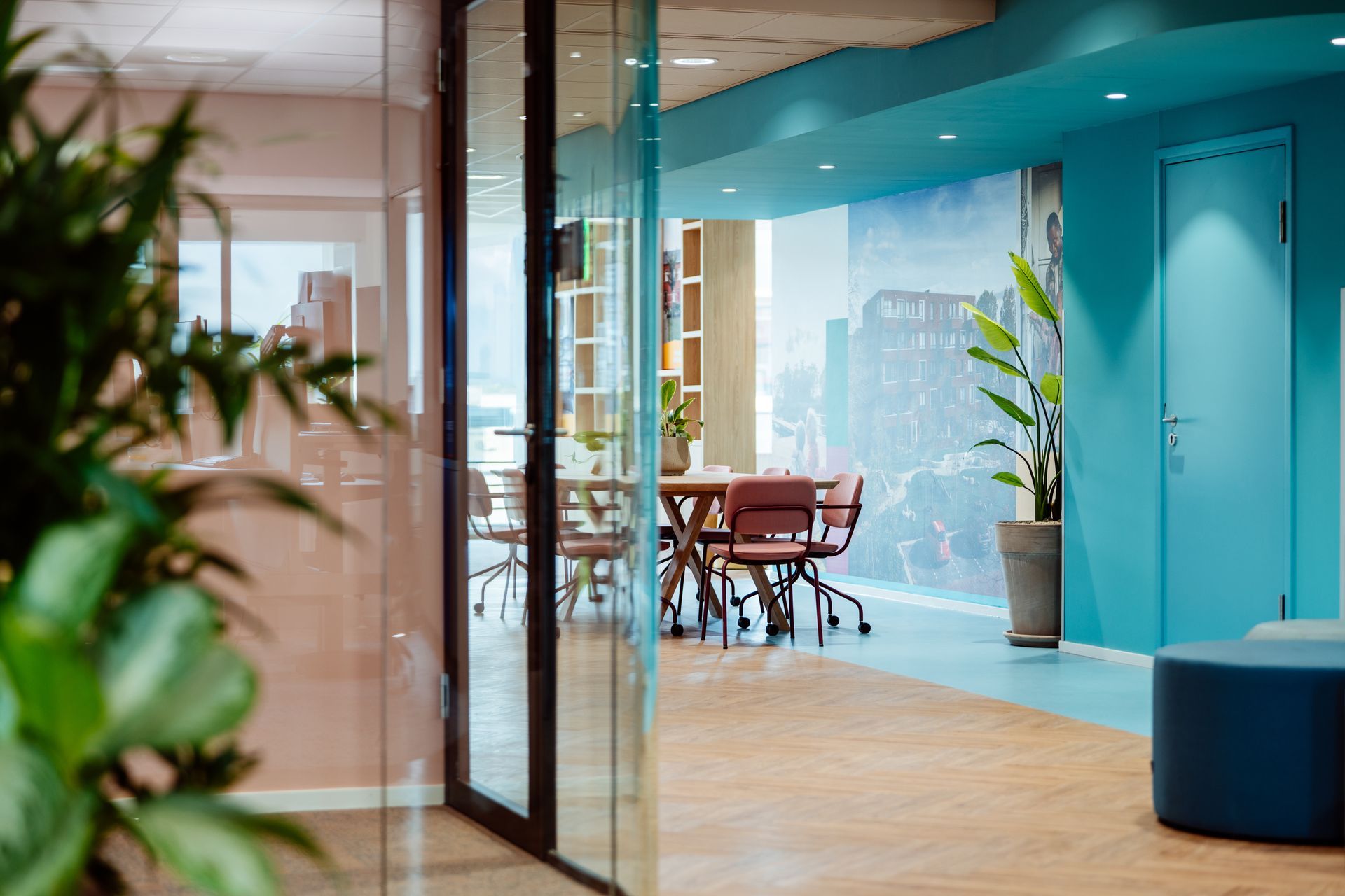 Moderne kantoorlounge met turquoise muren, houten vloeren, glazen scheidingswanden en een vergadertafel met stoelen.
