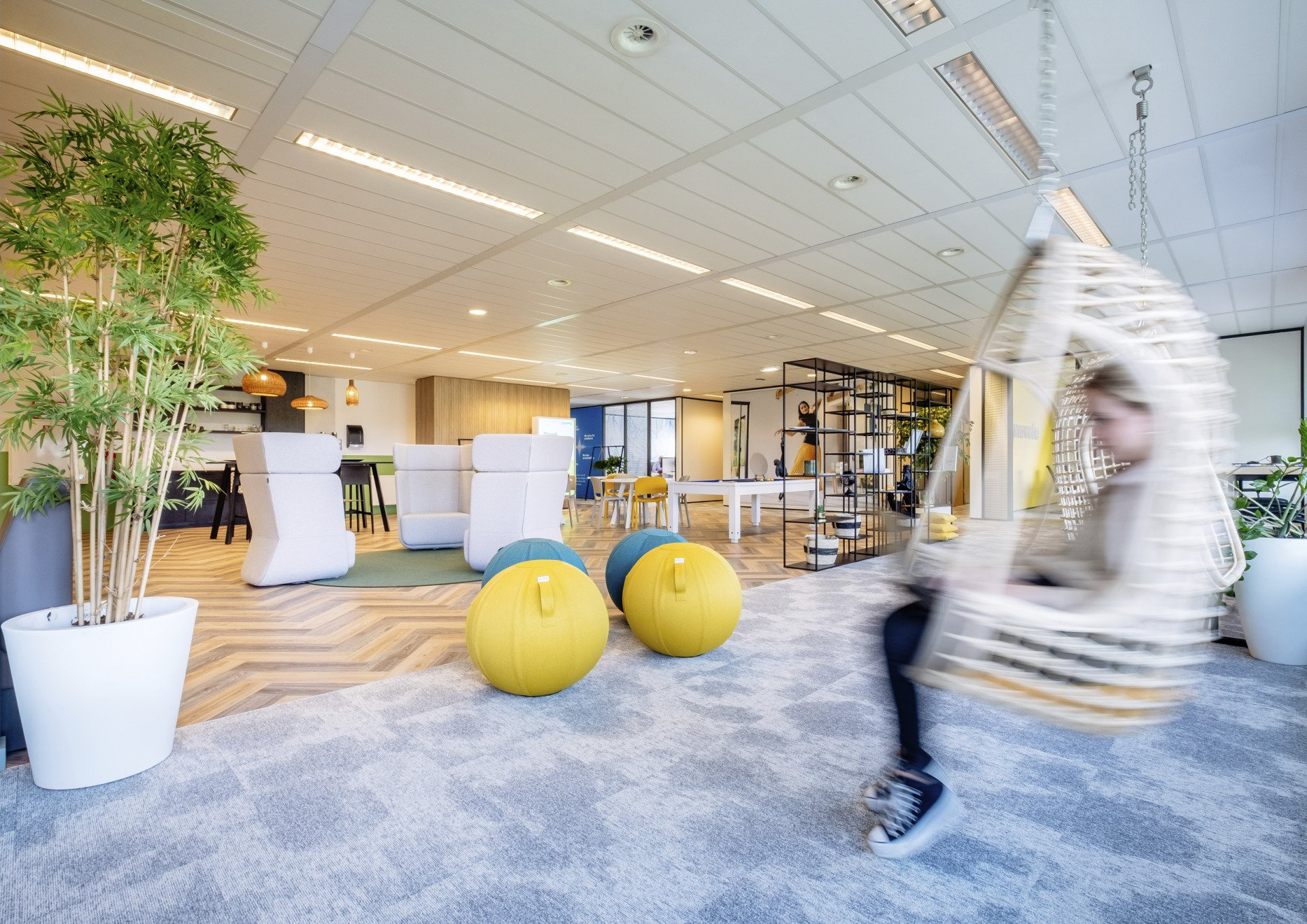 Lichte kantoorlounge met gele ballen, planten en een wazige figuur die voorbijloopt.