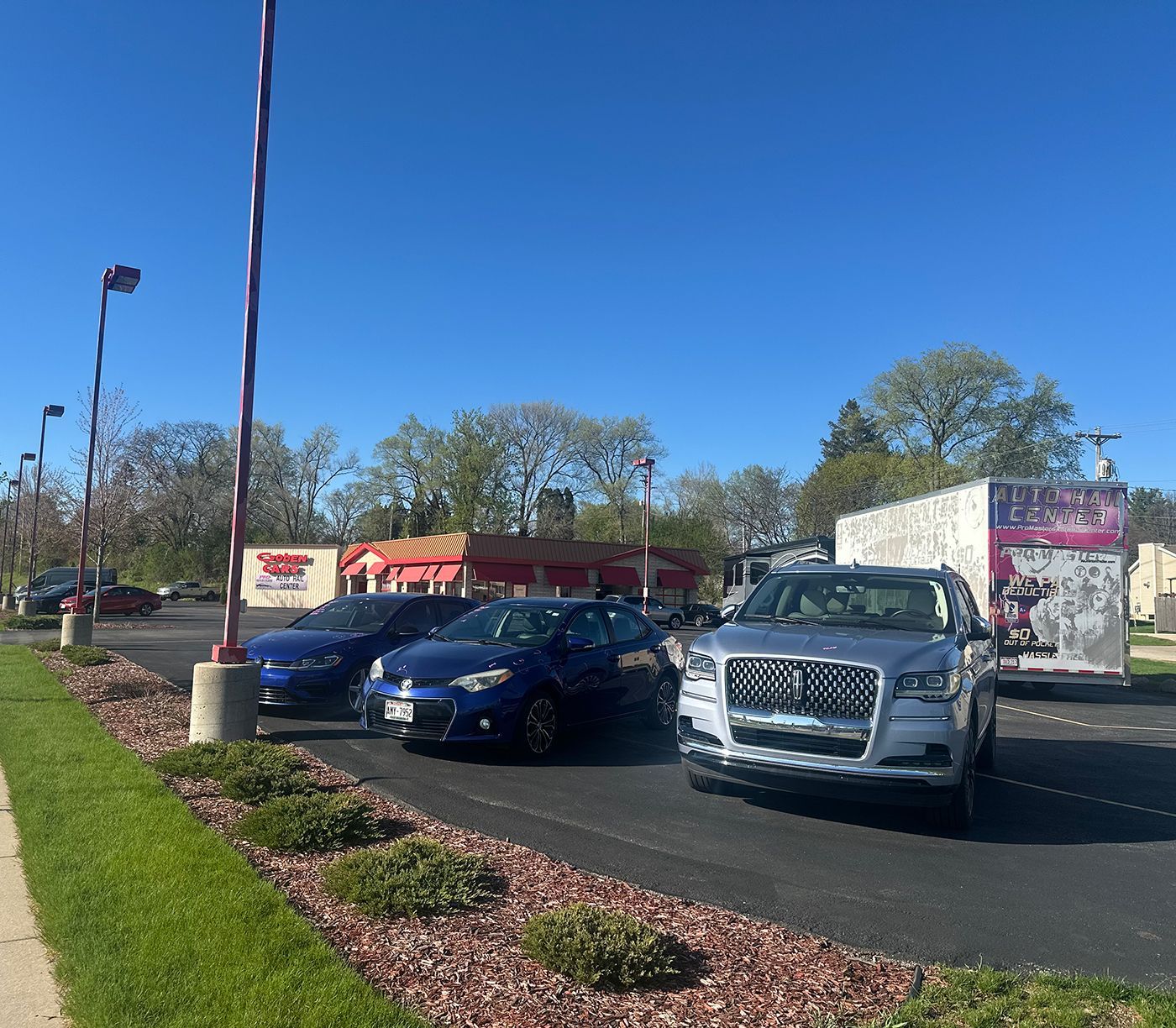 Parking lot with a blue car and silver SUV near a sign under a clear blue sky.