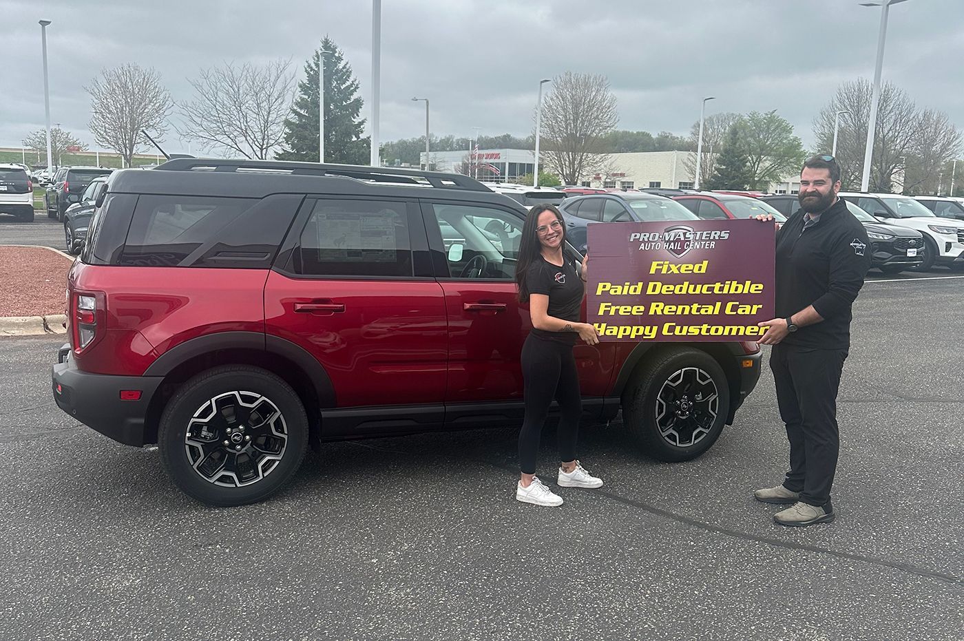 Two people posing beside a red SUV in a parking lot, holding a large sign.