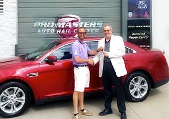 Two men are shaking hands in front of a red car.