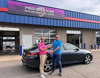 Two men are shaking hands in front of a car.