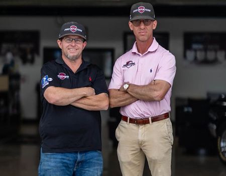 Two men stand with arms crossed in a garage, wearing branded caps and casual shirts.