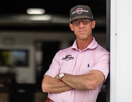 Man in pink shirt and black cap standing with arms crossed in a garage doorway