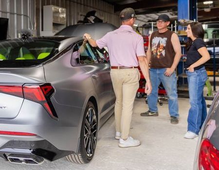 Three people examine a silver sports car in a garage showroom