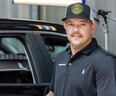 Man in black polo and cap standing beside a black truck in a garage.