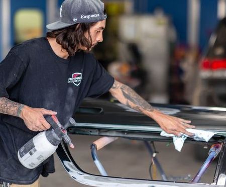 Mechanic sprays a car door panel in a workshop, wearing a cap and black shirt.