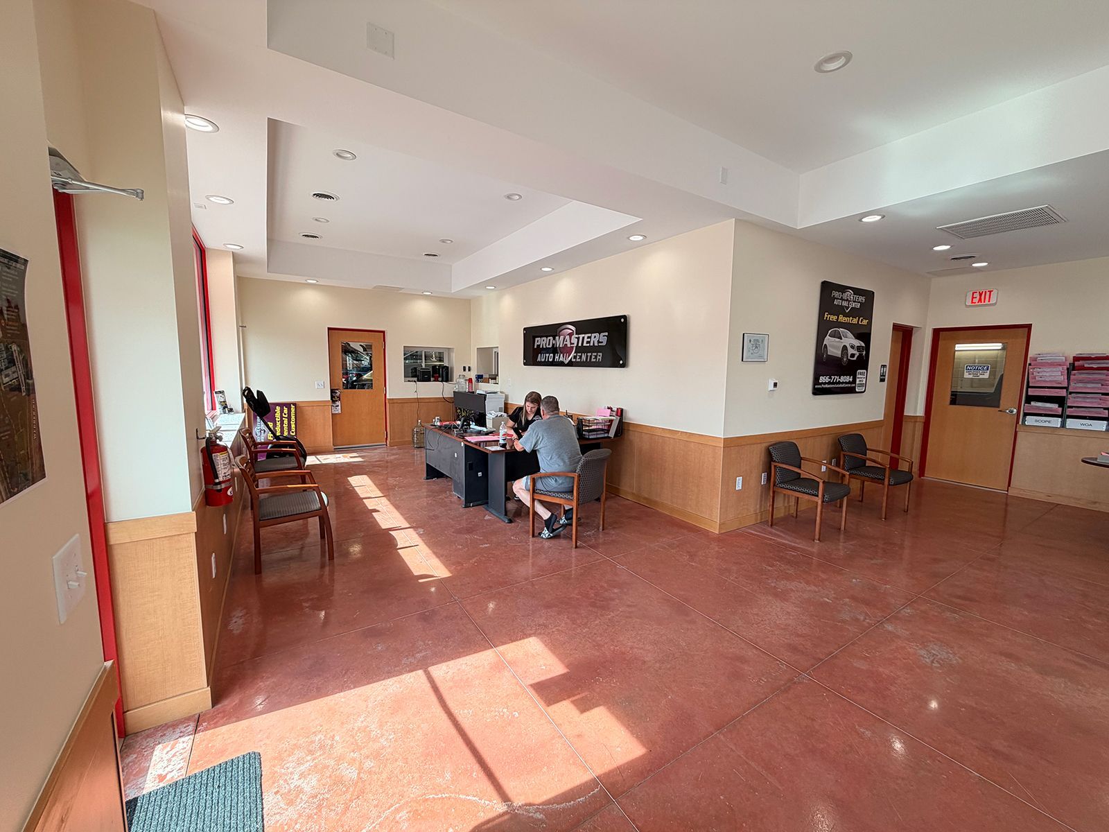 Bright lobby with red tile floor, sunlight, tables, chairs, and a few people seated near a service counter.