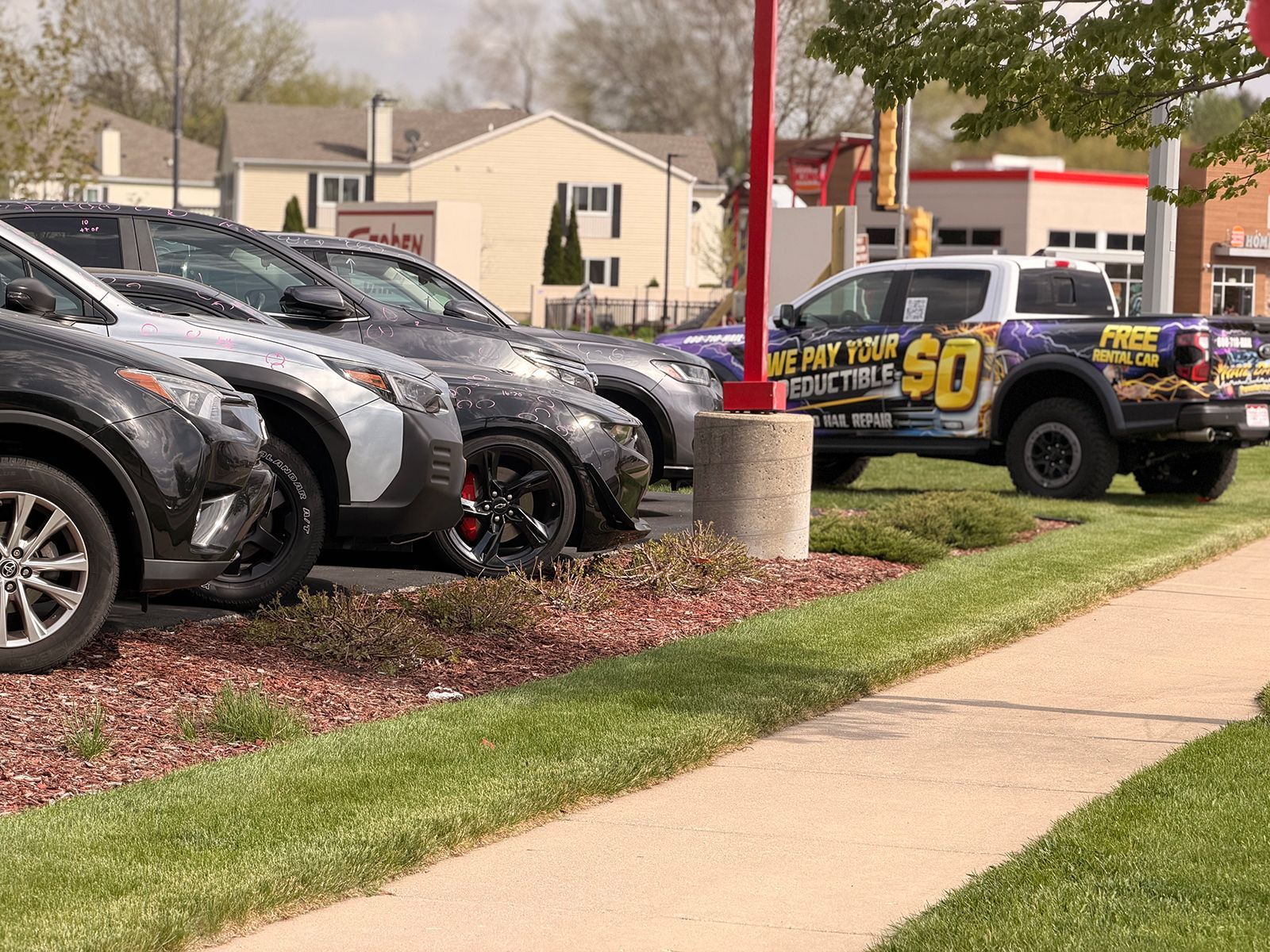 Dealer lot with parked SUVs and a black-and-yellow race truck on a grassy edge by a sidewalk
