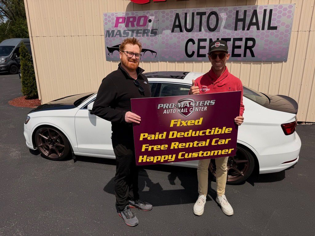 Two people hold a sign in front of an auto hail center, standing by a white convertible car.