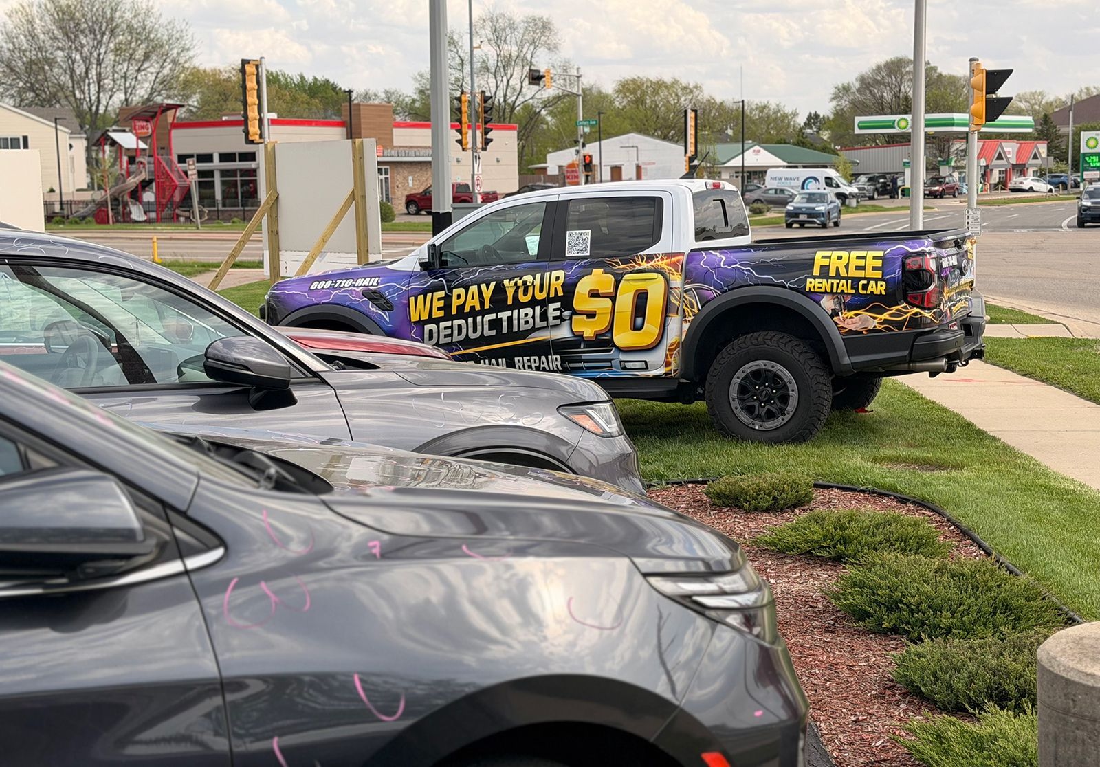 Wrapped pickup truck in a parking lot advertising “FREE” and “We pay your deductible” beside parked cars