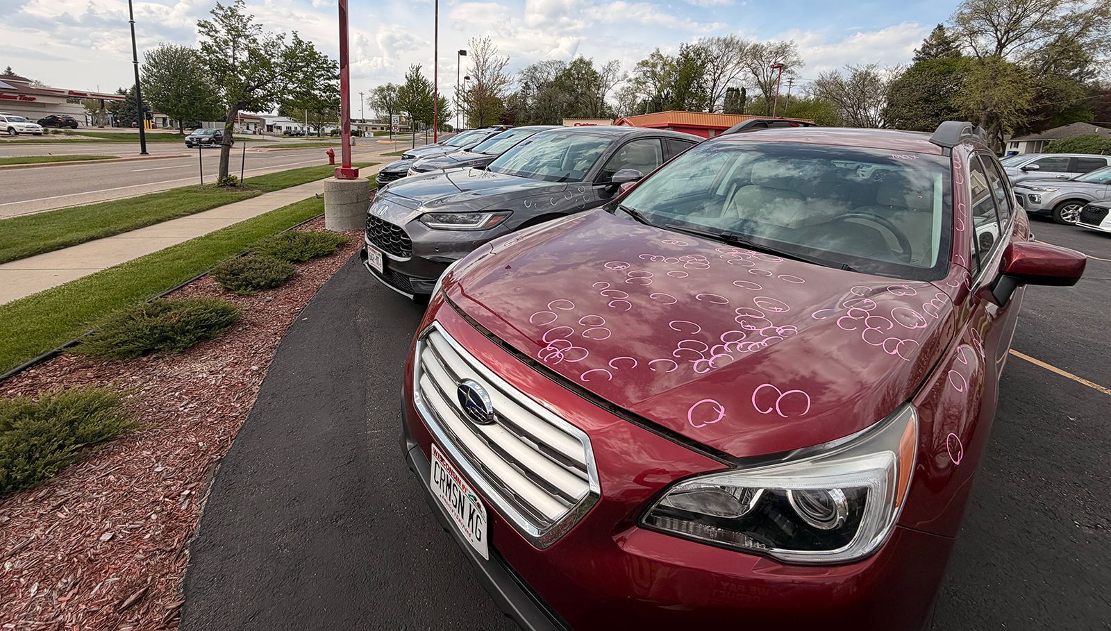 Maroon sedan and dark gray car parked on a wet dealership lot under cloudy skies