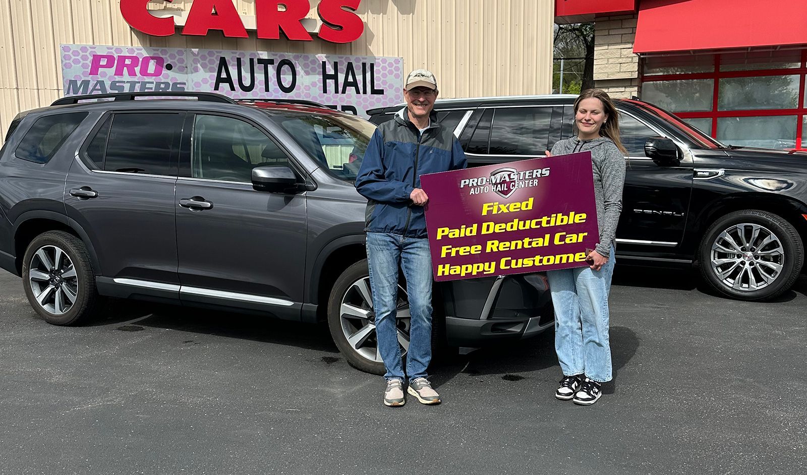 Two people holding a sign in front of a dark SUV outside an auto hail repair shop