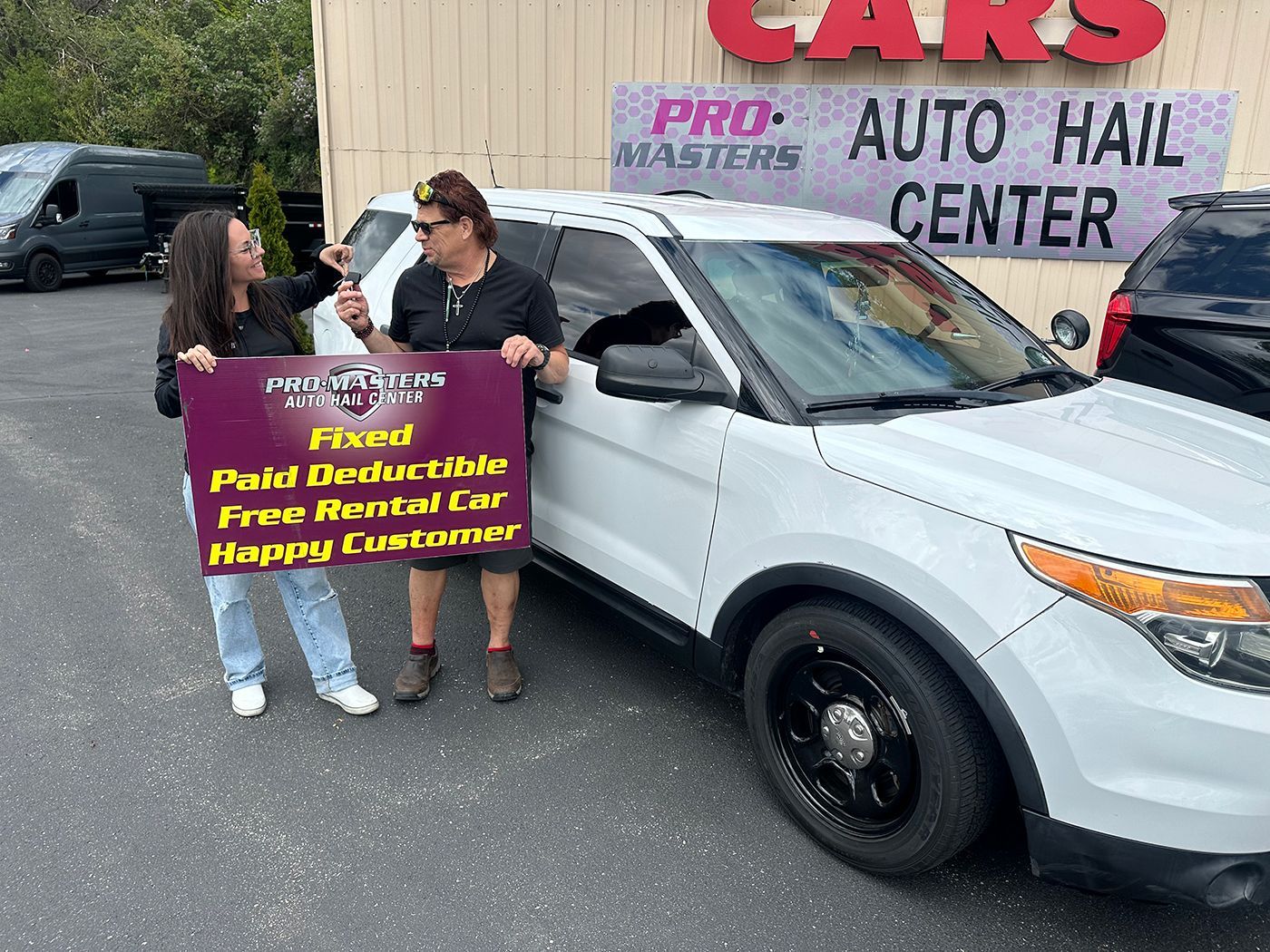 Two people hold a protest sign beside a white SUV outside a car center.