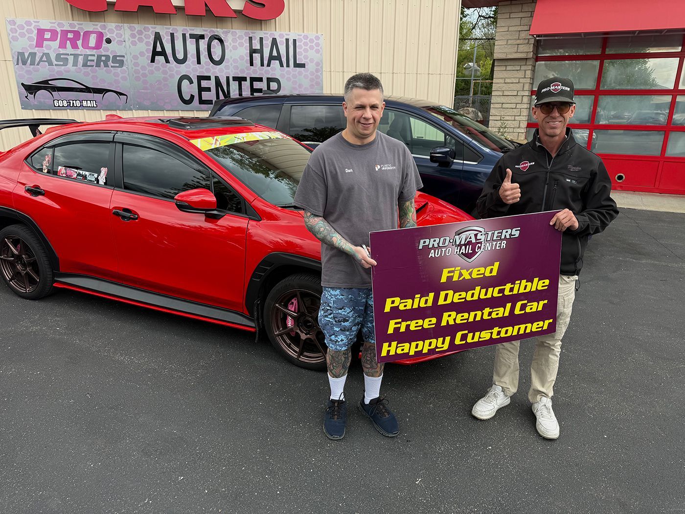 Two men hold a sign in front of a red car at an auto hail center.