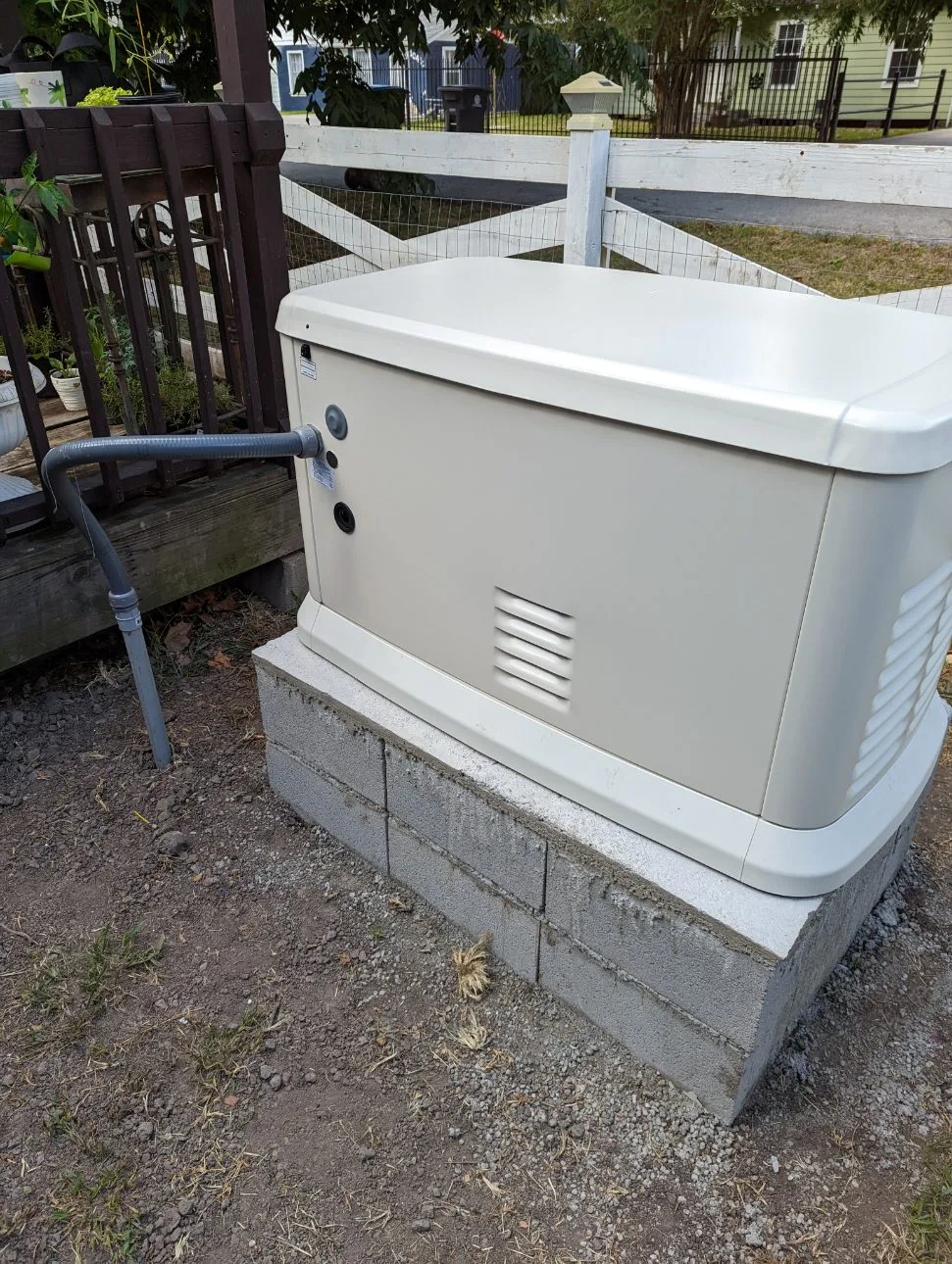 A generator sits on cinder blocks near a fence. The beige machine has a vent and is connected to a metal pipe.