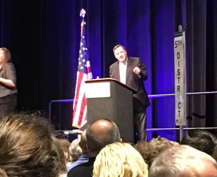 A man in a suit speaks at a podium with an American flag.
