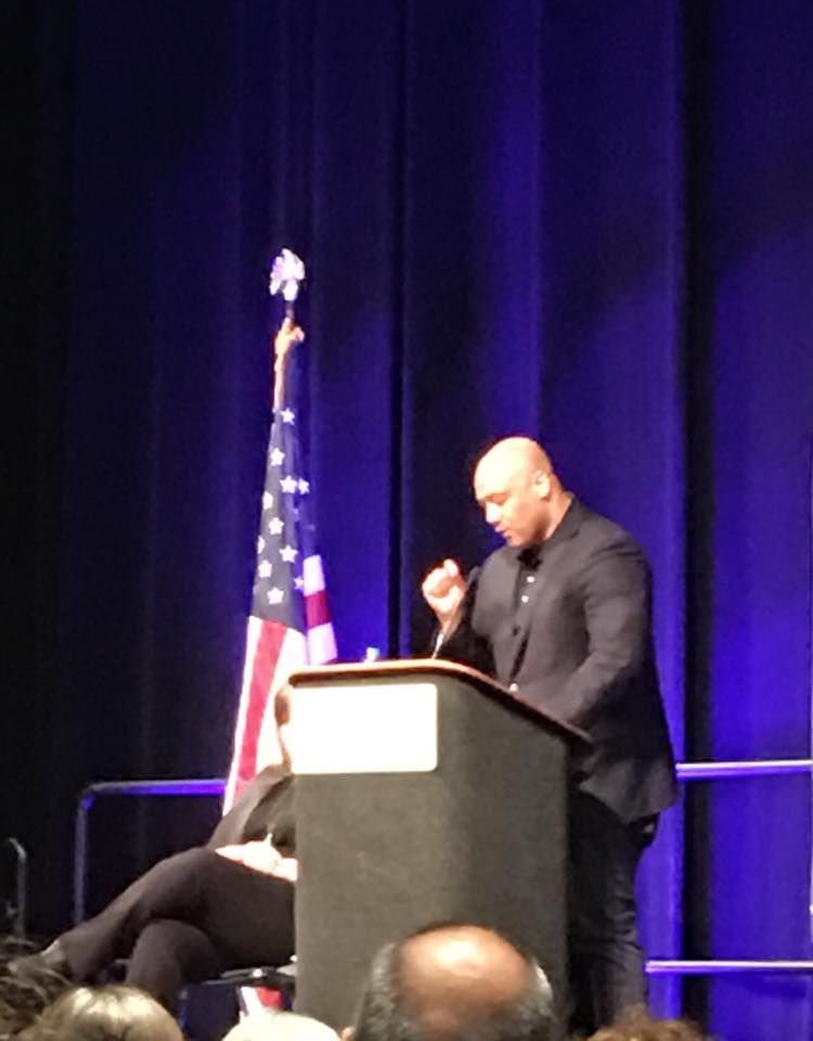 A man in a black suit speaks at a podium with the American flag in the background, addressing an audience.