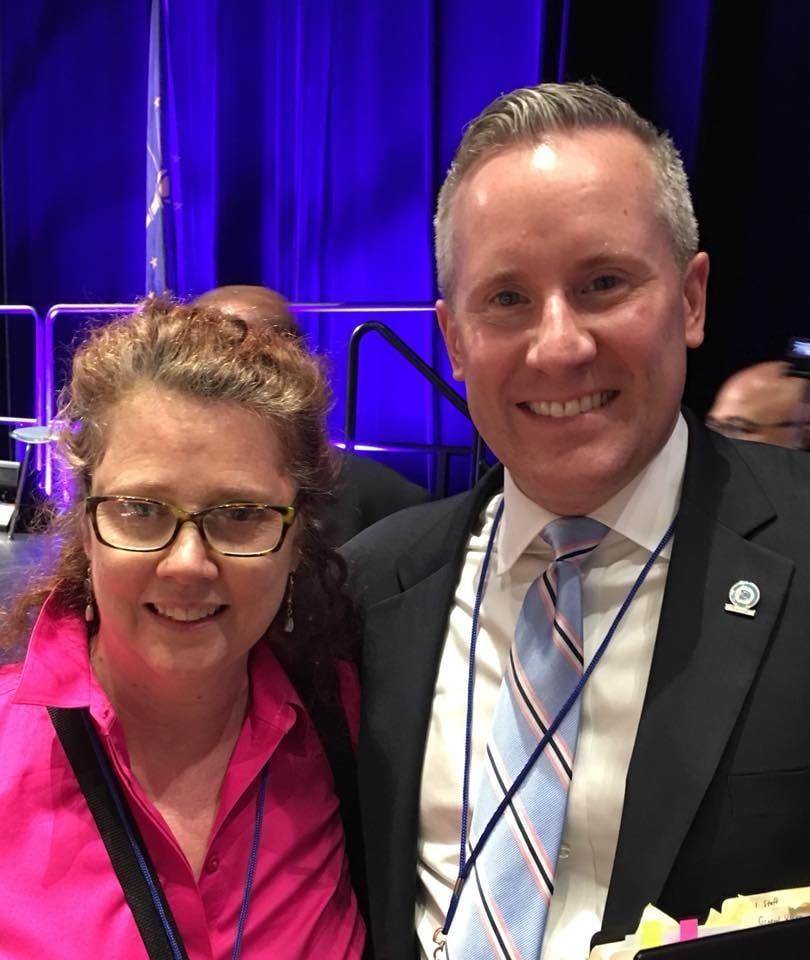 A woman in glasses and a pink shirt smiles next to a man in a suit and tie.