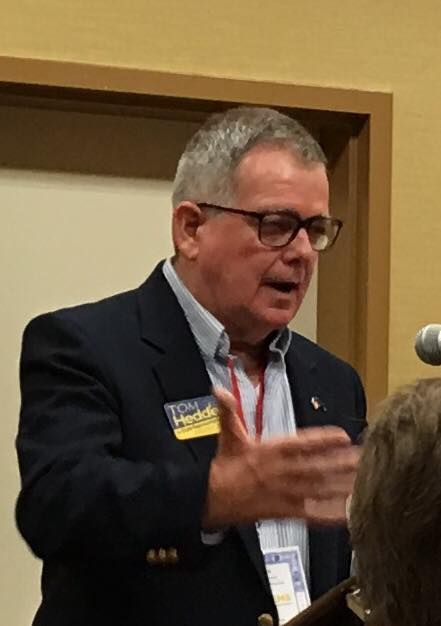 Man in a blue blazer and glasses speaking, gesturing with his right hand.