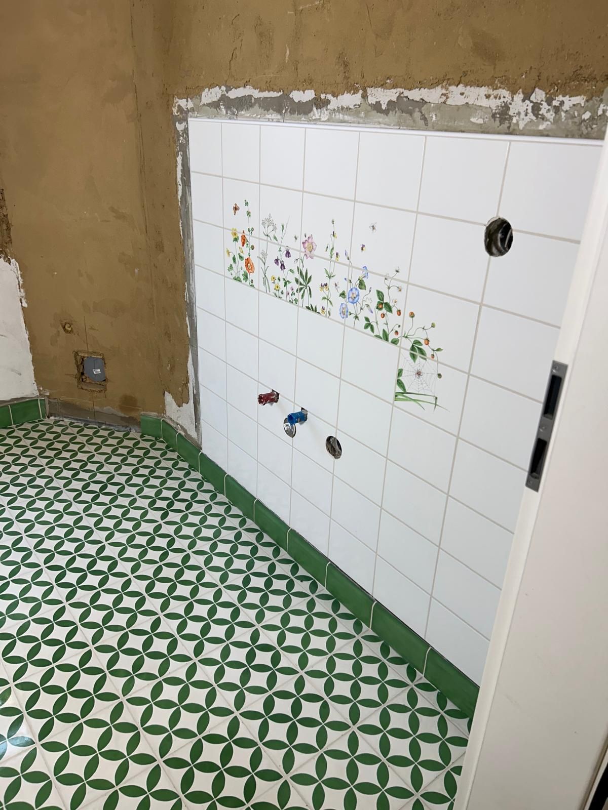 Wand mit verlegten weißen Fliesen, darunter ein dekoratives Blumenmotiv; darüber unverputzte Fläche im Rohbauzustand