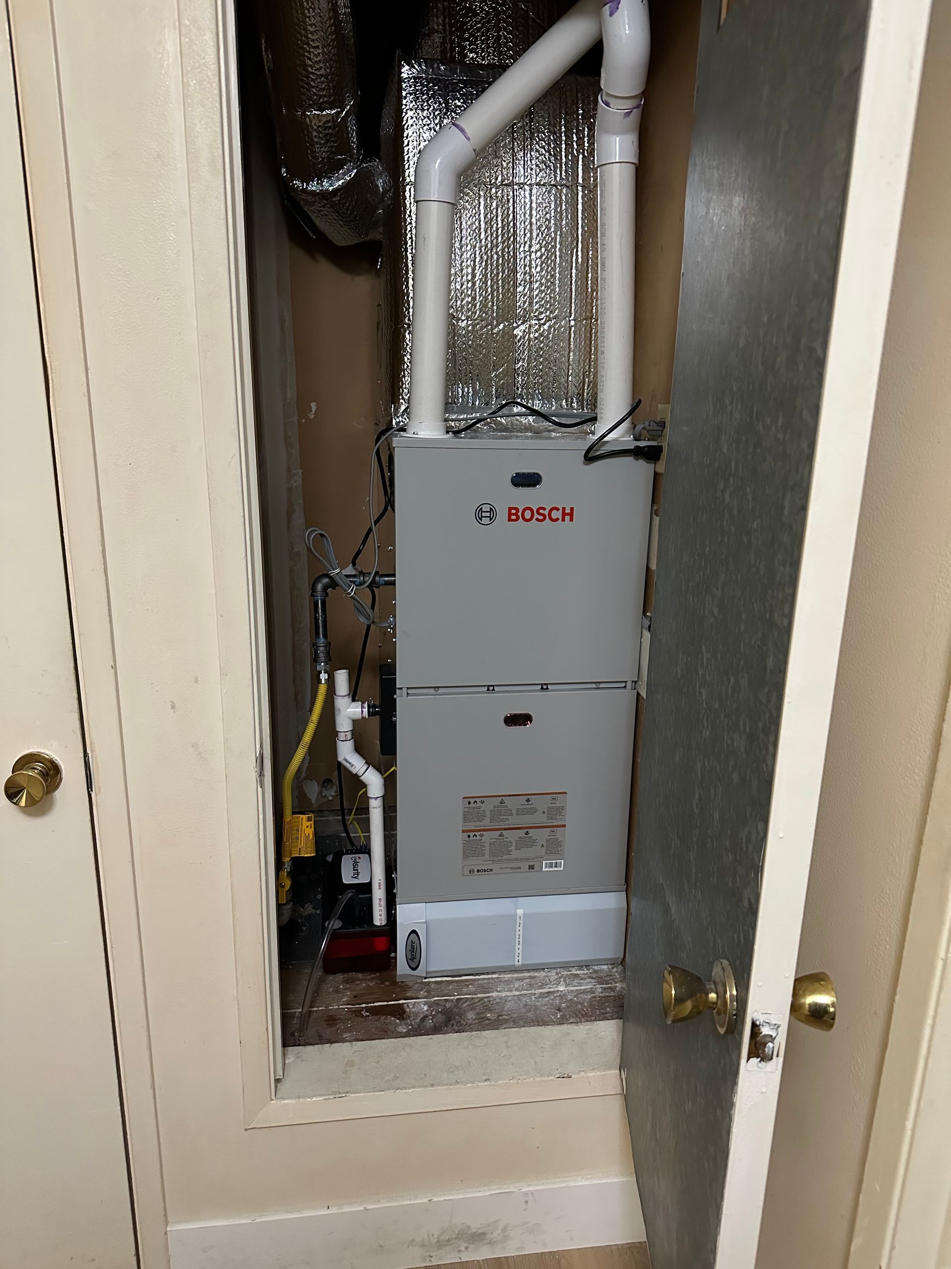 A Bosch furnace installed in a closet, with white vent pipes and a yellow gas line visible. The closet walls are beige.