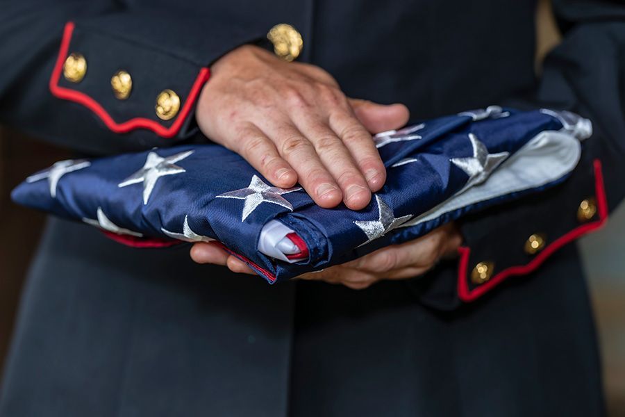 Hands holding a folded American flag. The flag is blue with white stars, red and white stripes. Person in military uniform.