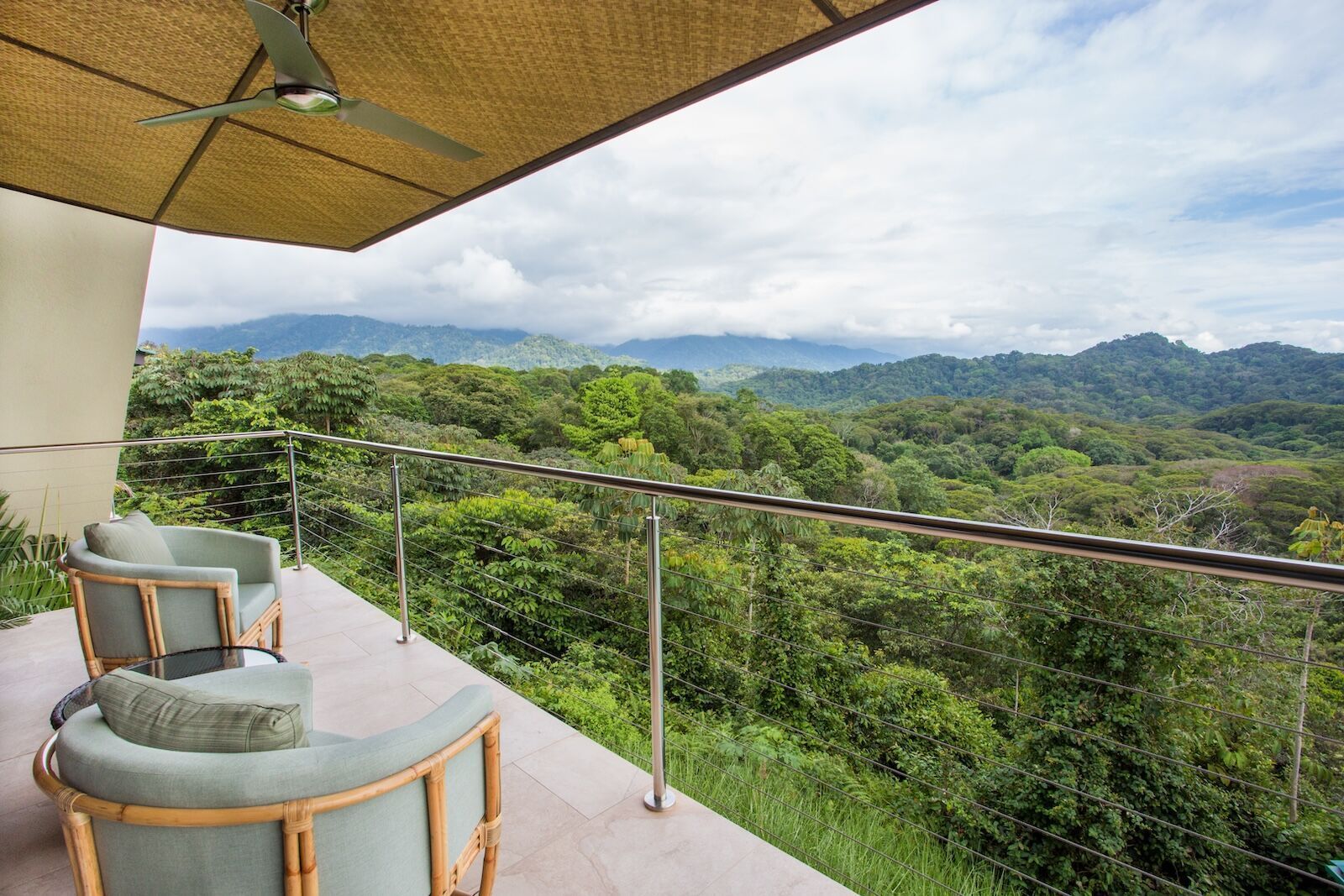 A balcony with chairs and a ceiling fan overlooking a lush green forest.