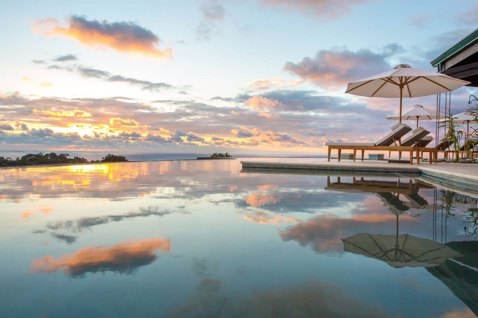 A swimming pool with chairs and umbrellas and a view of the ocean at sunset.