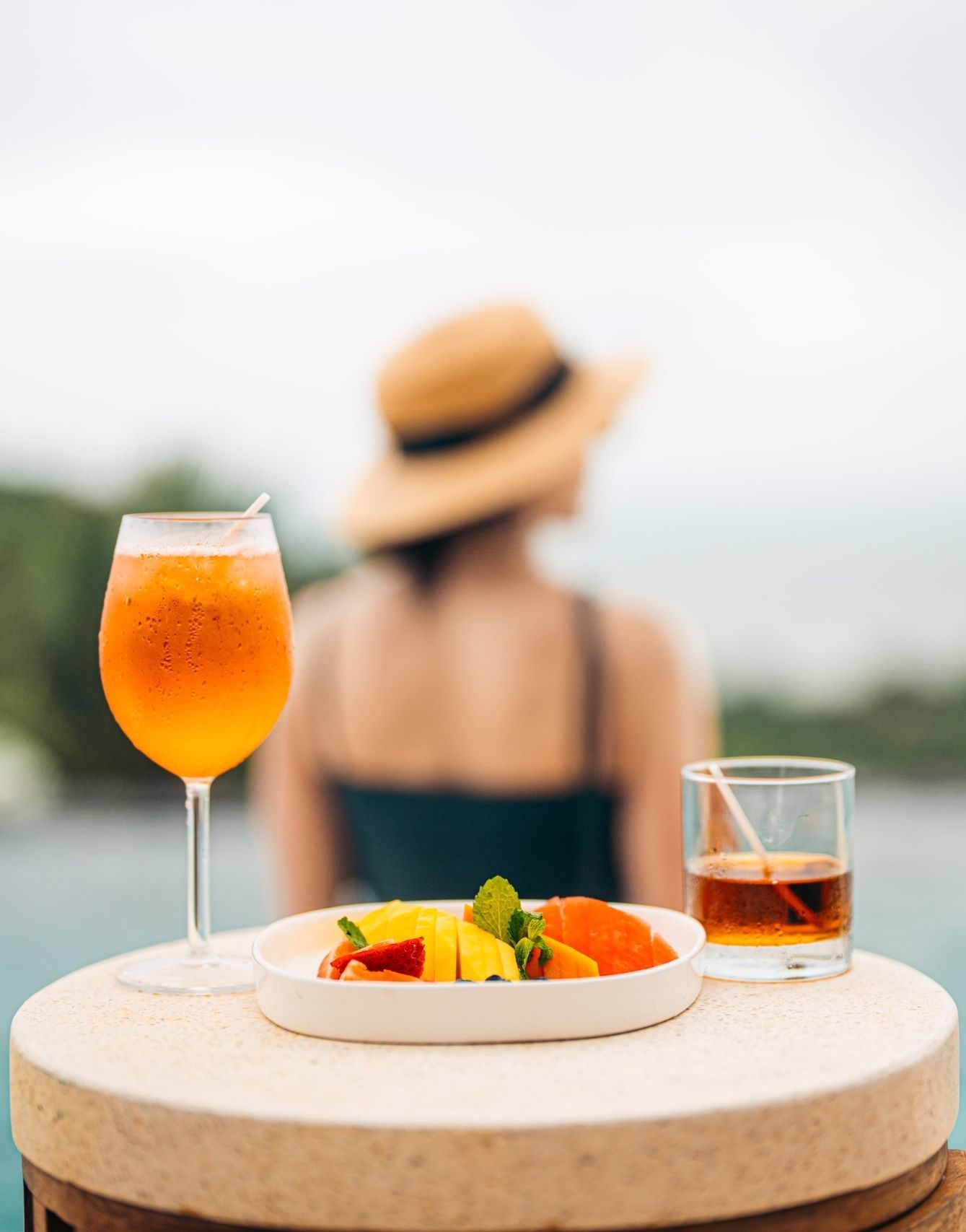 A woman in a straw hat sits behind a plate of fruit and a glass of wine