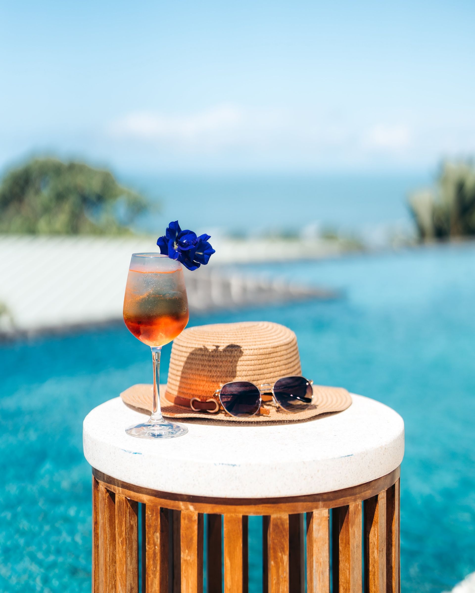 A straw hat , sunglasses , and a glass of wine are on a table next to a swimming pool.
