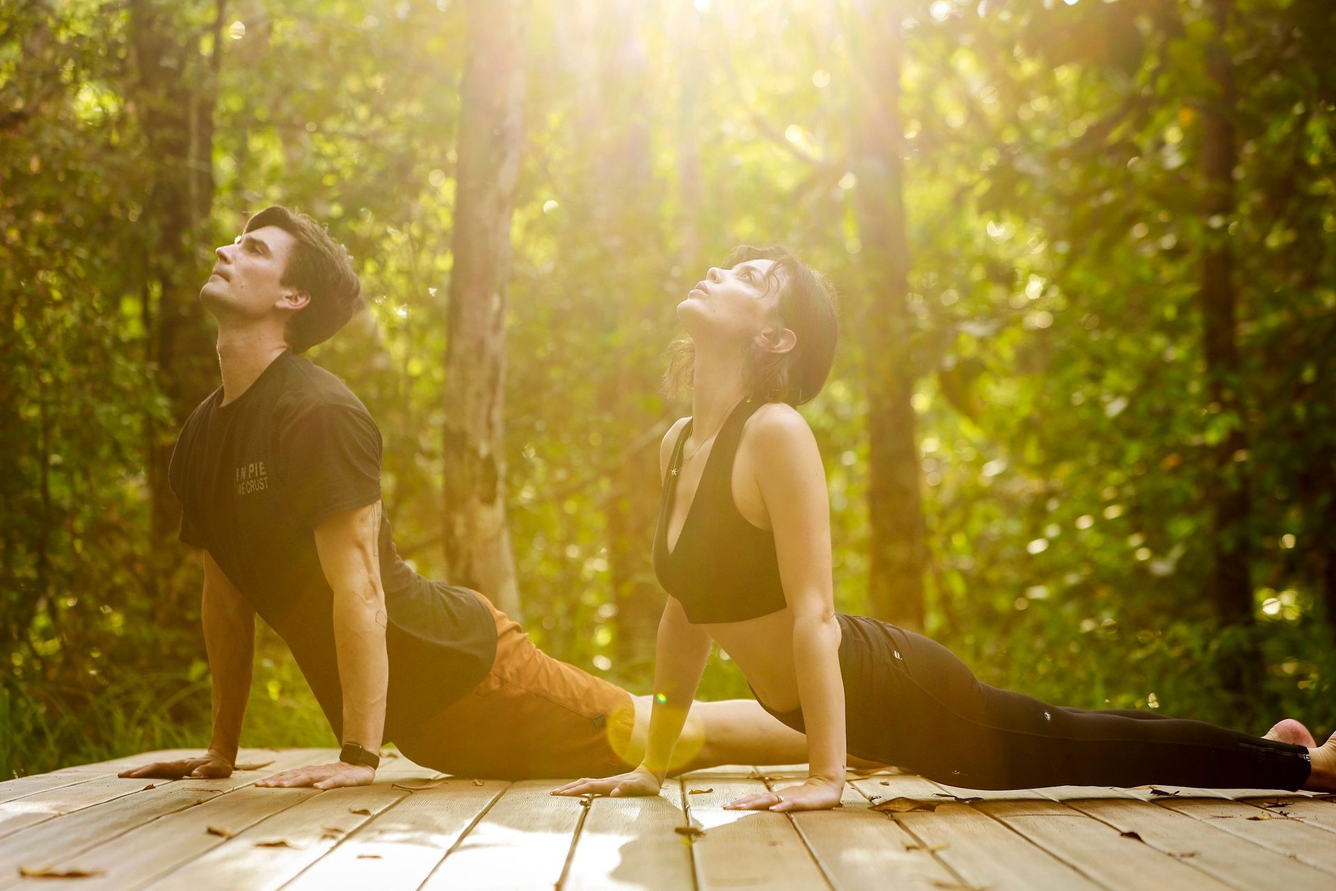 Two people doing yoga in a sunlit forest, looking up in a cobra pose.