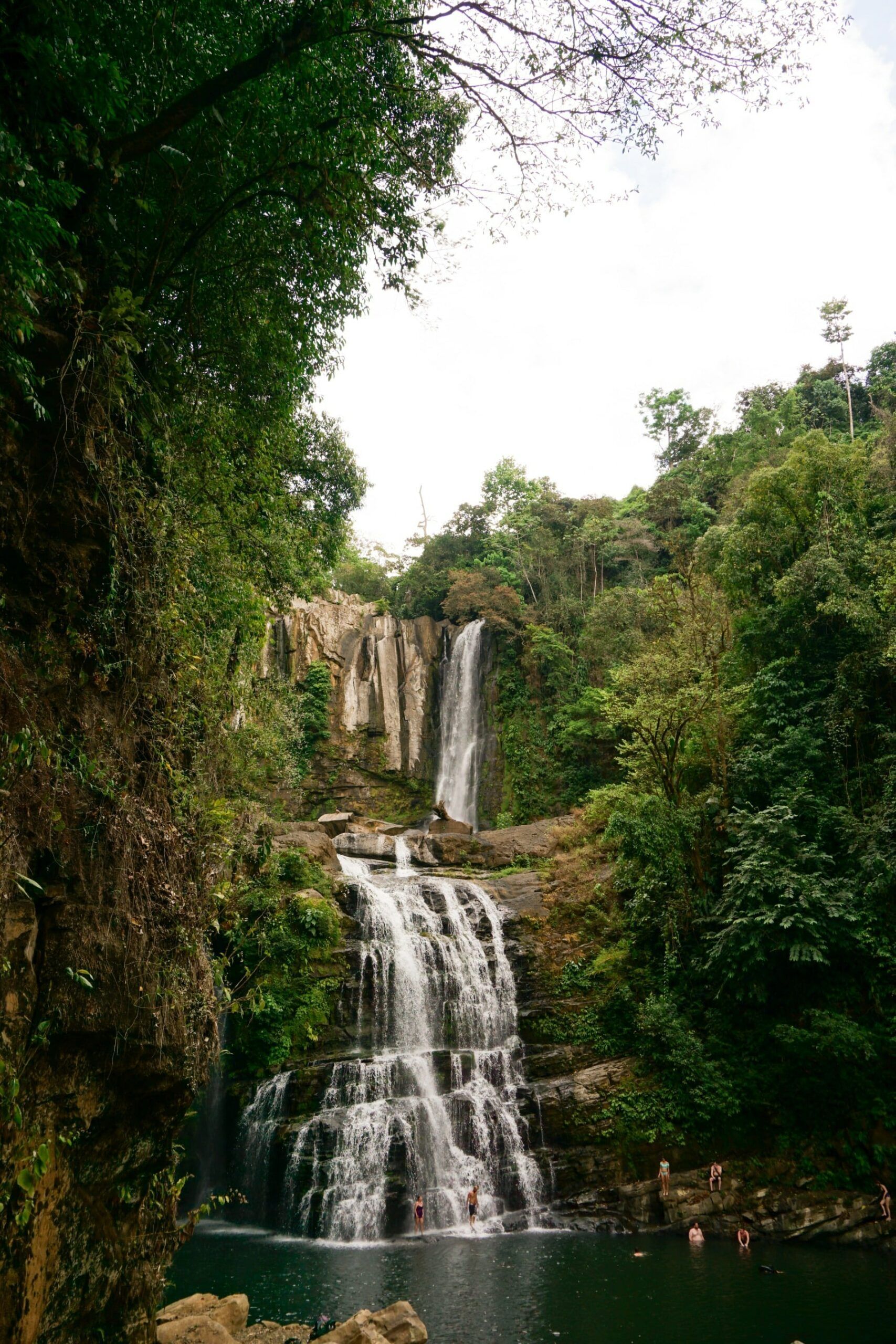 Waterfall cascading into a pool surrounded by lush green foliage; some people swimming.