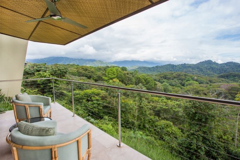 A balcony with chairs and a ceiling fan overlooking a lush green forest.