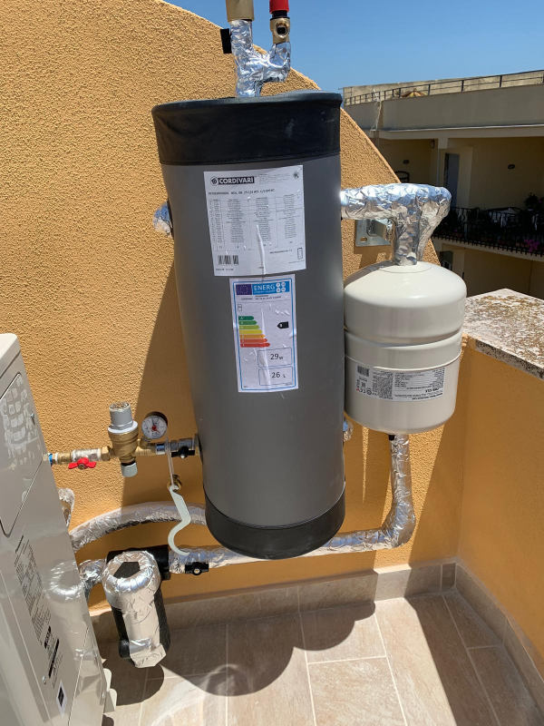 Uno scaldabagno cilindrico grigio con vaso di espansione bianco annesso, montato su una parete esterna beige e soleggiata.