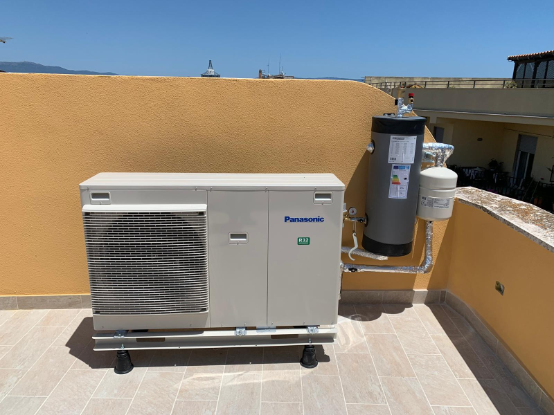 Un'unità pompa di calore Panasonic e un accumulatore di acqua calda installati su una terrazza residenziale con pareti color beige e pavimento piastrellato.
