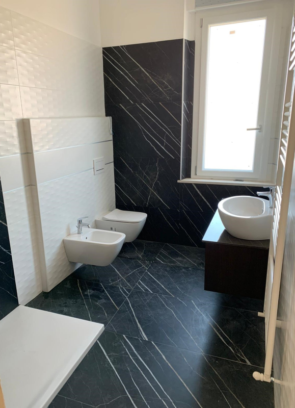 Bagno moderno con pavimenti e pareti in marmo venato nero, bidet bianco, WC, lavabo con mobiletto e piatto doccia.